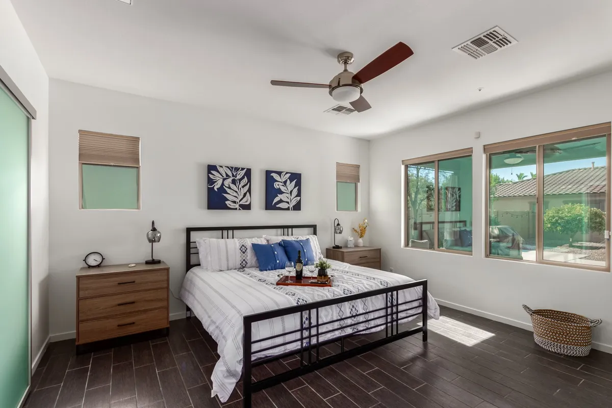 A tranquil bedroom scene with a plush bed adorned with decorative pillows, framed artwork on the wall, natural light streaming through large windows, and a cozy nightstand with a lamp. The setting exudes comfort and relaxation, perfect for unwinding at the end of the day.