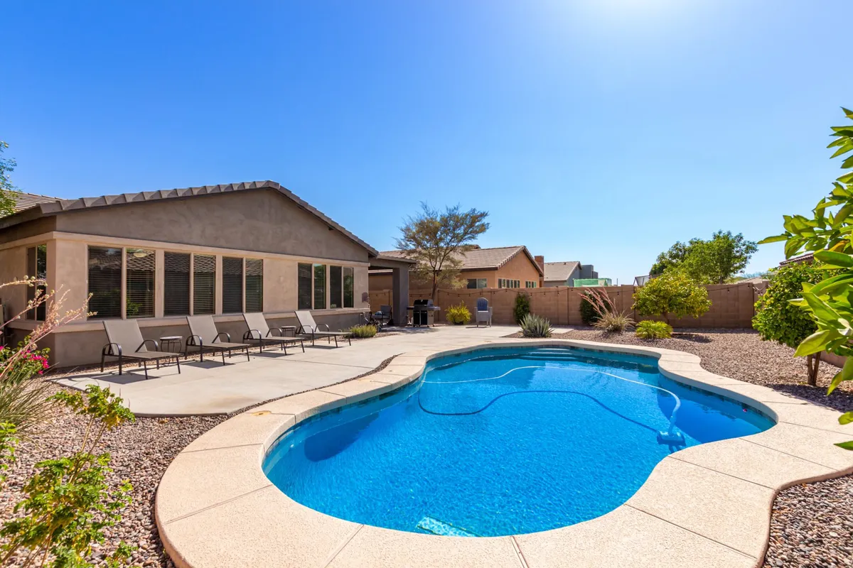An expansive backyard features a sparkling swimming pool, a shaded patio area with comfortable seating, and landscape plants that enhance privacy. The space is designed for both sunbathing and outdoor dining, inviting mornings with coffee or outdoor meals in a peaceful, fenced setting.