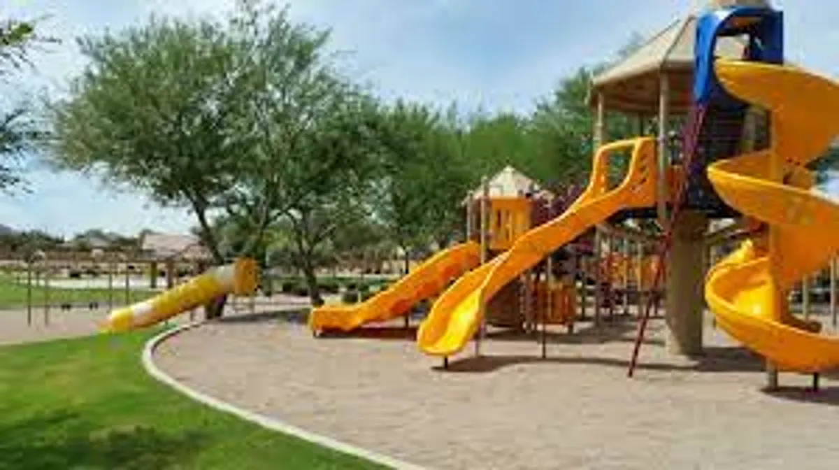 A vibrant playground scene featuring colorful slides, swings, green grass, and mature trees. Children can be seen playing and families gathering for picnics beneath the shade. The environment is lively and welcoming, capturing a sense of community and outdoor fun.
