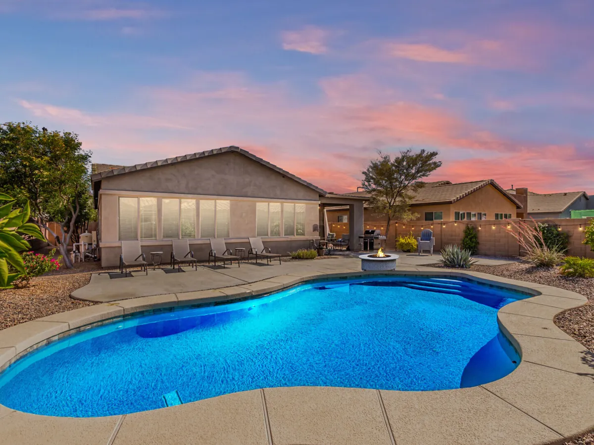 A sparkling swimming pool with inviting blue water is surrounded by a landscaped backyard. Four sun loungers are arranged on the pool deck, and a fire pit with comfortable outdoor seating provides an ideal space for relaxing or socializing. Lush greenery and palm trees frame the space, creating a tranquil, resort-like environment perfect for enjoying the Arizona sunshine.