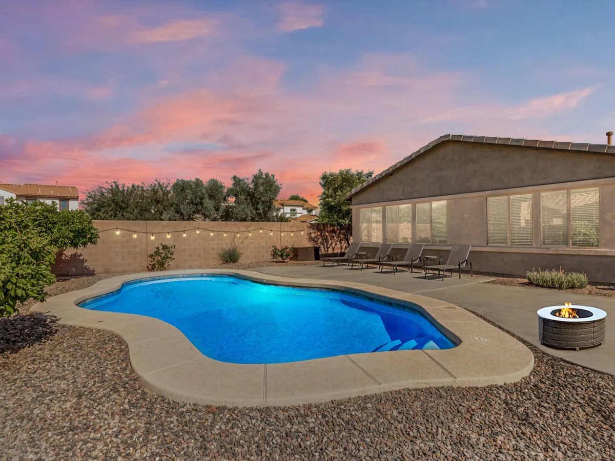 A sunlit backyard with a sparkling swimming pool, surrounded by lounge chairs and landscaped greenery. The scene includes a fire pit area, perfect for evening gatherings, and a casual, social atmosphere ideal for family poolside relaxation.