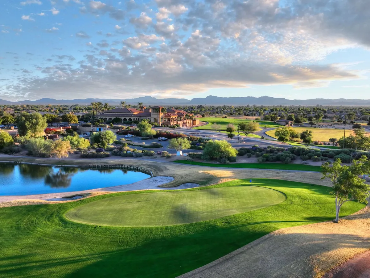 A wide shot of the Seville community's lush golf course, featuring a sparkling pond, the inviting clubhouse, and scenic mountains in the background. The greens are well-manicured and the outdoor atmosphere is relaxed, with space for walking, picnics, and community gatherings.